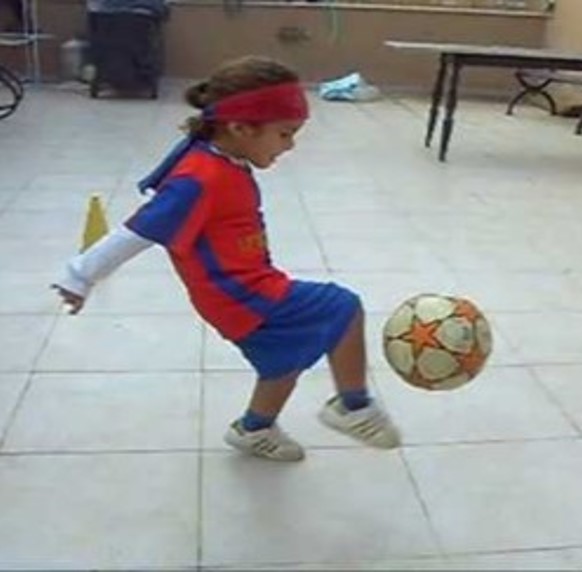 This photo shows a youngster kicking a soccer ball. The child’s arms are held backward while the head is tipped forward.