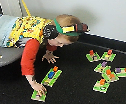 this photo shows a child who is prone on a platform swing. He is playing a game with wooden car & truck dominoes. The vehicles are posed in different spatial orientations.