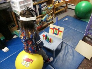 this photo shows a child who is sitting on a physioball while engaging spatial orientation of building a tower of blocks. The back is extended while the head is straight upright.