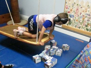 this is a photo of a boy who is quadruped on a platform swing. His upper back and neck are extended as he looks down at a pile of puzzle cubes that he will be working to assemble.